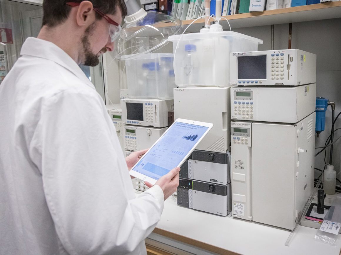 Scientist in a lab coat and safety glasses analyzing data on a tablet next to analytical laboratory equipment including HPLC or chromatography systems.